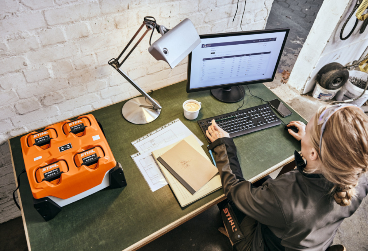 Απεικονίζεται μια γυναίκα να εργάζεται στον υπολογιστή όσο δίπλα φορτίζουν 4 μπαταρίες στον πολυφορτιστή.